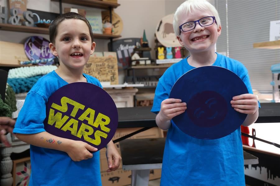 Two boys smile to the camera while holding up their art projects depicting the star wars logo and a smiley face.